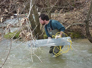Una imagen de un biólogo colocando una gran estructura de alambre en el agua que se conoce como muestreador químico pasivo.