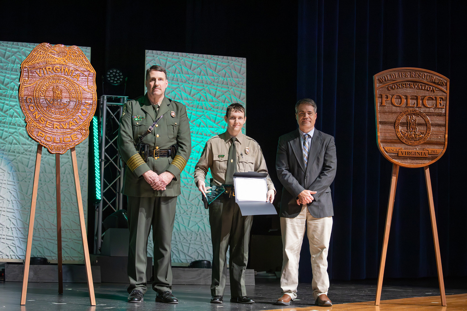 Una foto de un agente de la Policía de Conservación uniformado sosteniendo un premio de pie en un escenario entre otro CPO uniformado y un hombre con americana.