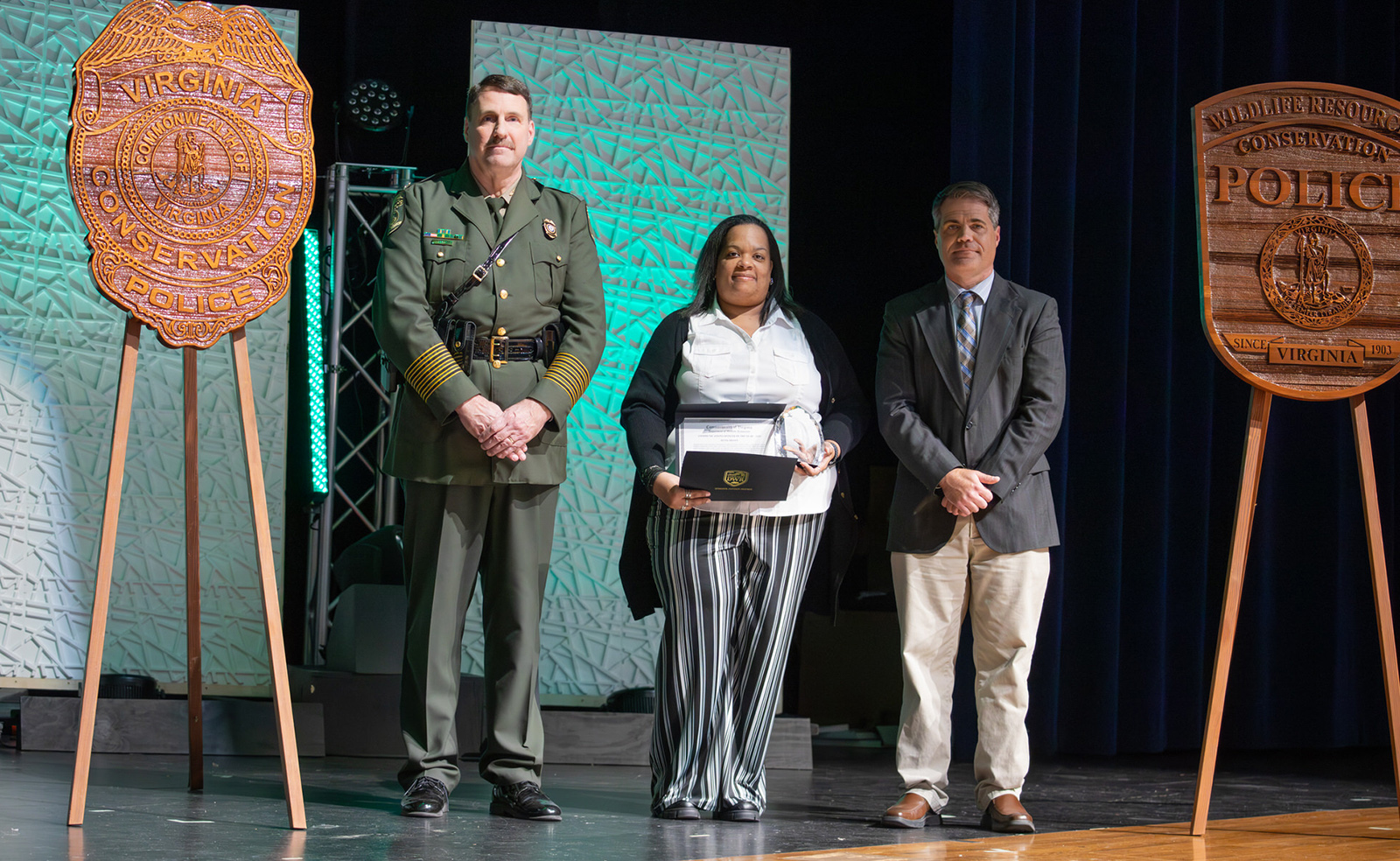 Una foto de una mujer sosteniendo un premio, de pie en un escenario con un hombre con uniforme de la Policía de Conservación y otro hombre.