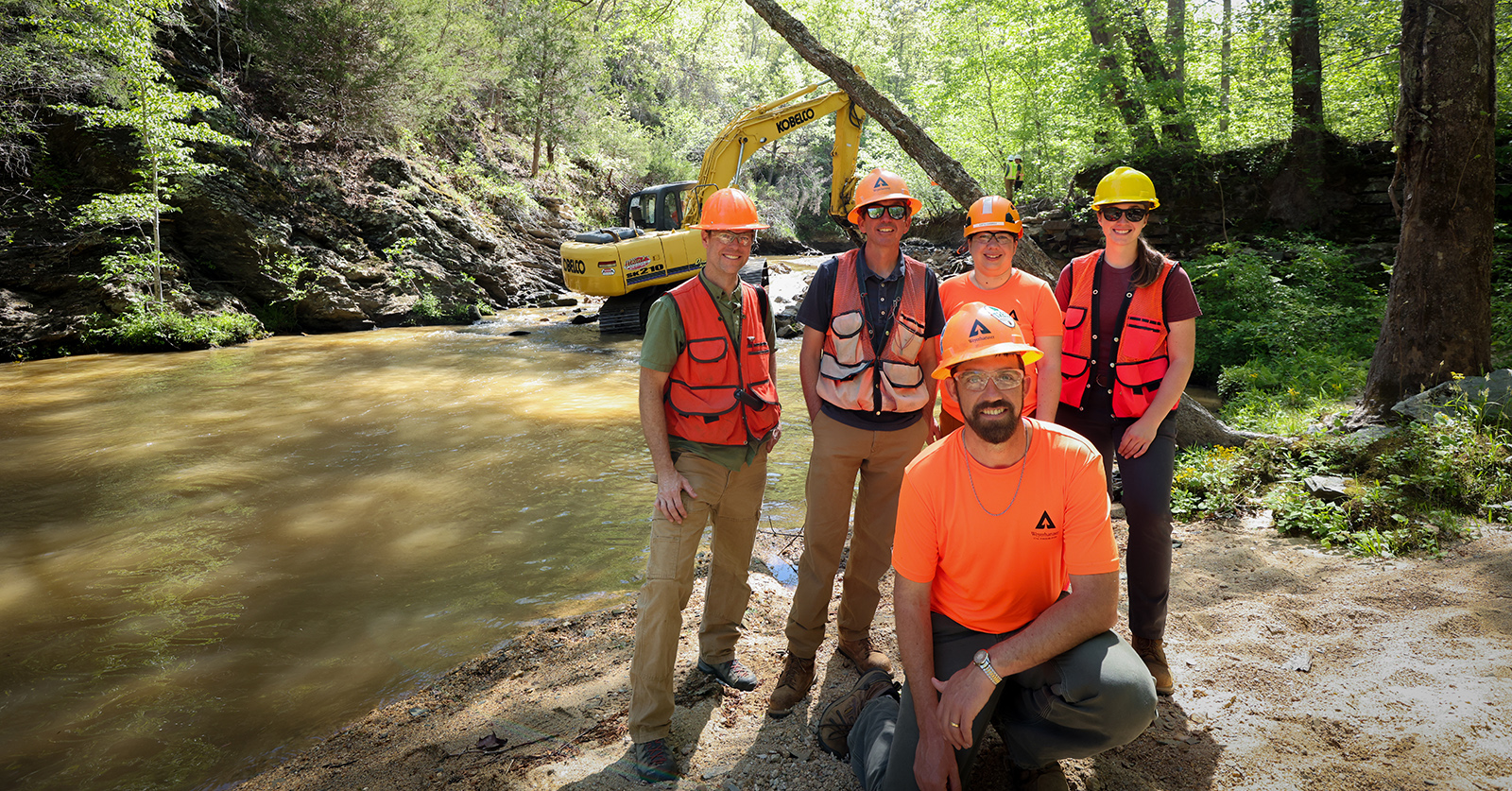Una foto de cinco personas con chalecos naranjas y cascos posando frente a una excavadora en un río poco profundo.
