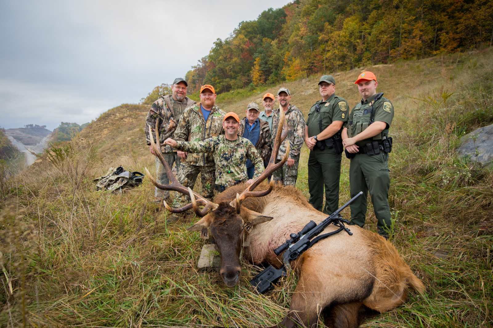 Una foto tomada de seis cazadores en camuflaje y naranja brillante posando con un gran alce macho tirado en el suelo y dos oficiales de la Policía de Conservación en uniforme.