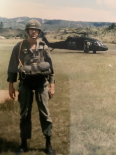 Una foto antigua de un hombre con uniforme militar de pie en un campo con un helicóptero Blackhawk en el fondo.