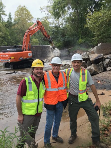 Construcción de remoción de la represa con un tractor retroexcavadora derribando la represa en la parte de atrás a la izquierda con tres hombres (el coordinador de Virginia Fish Passage, Alan Weaver, con socios de la agencia) de pie juntos sonriendo para una foto. 