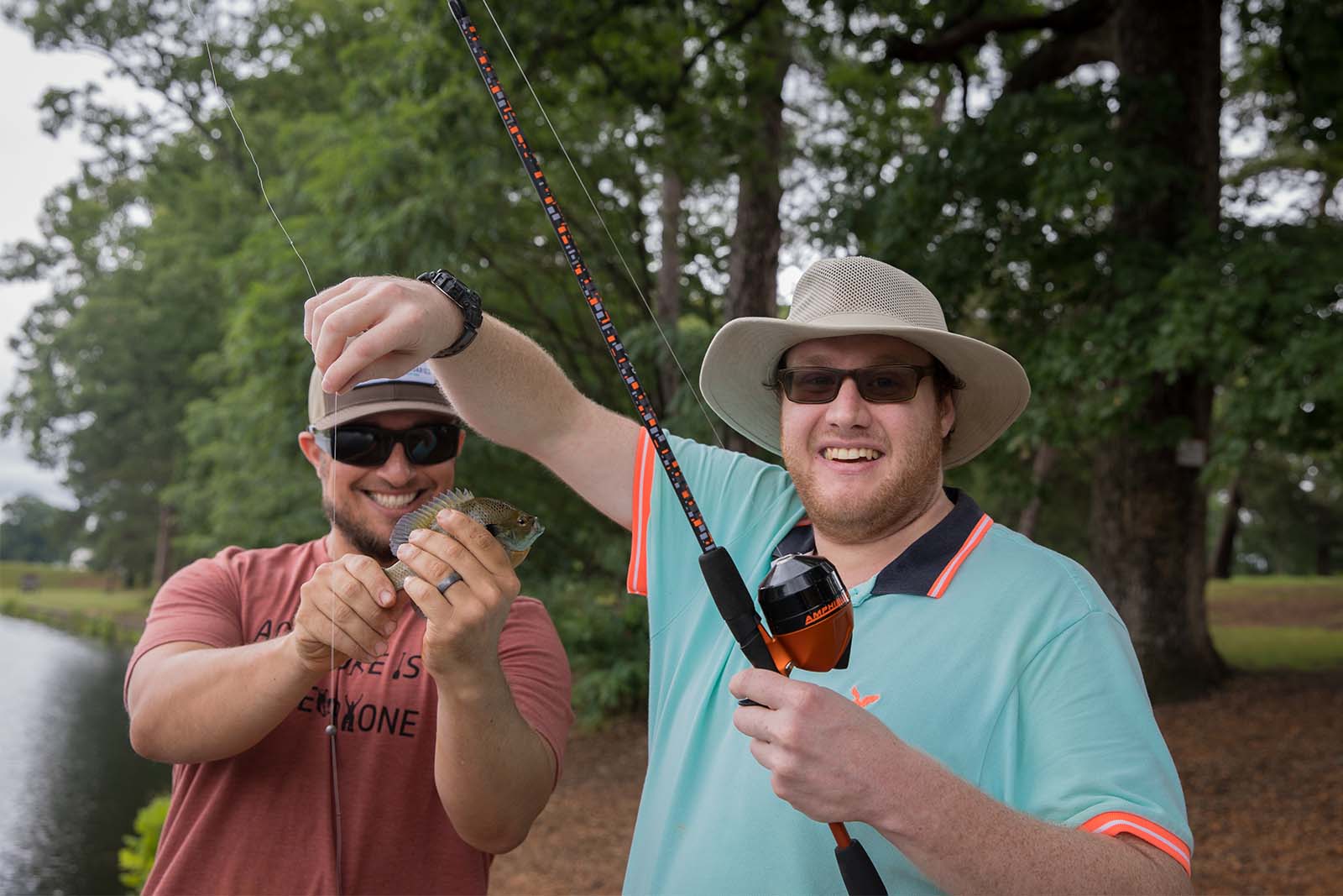 Dos hombres de pie en la orilla de un lago. Una persona sostiene una caña de pescar mientras que la otra sostiene un pez que se retiró del anzuelo
