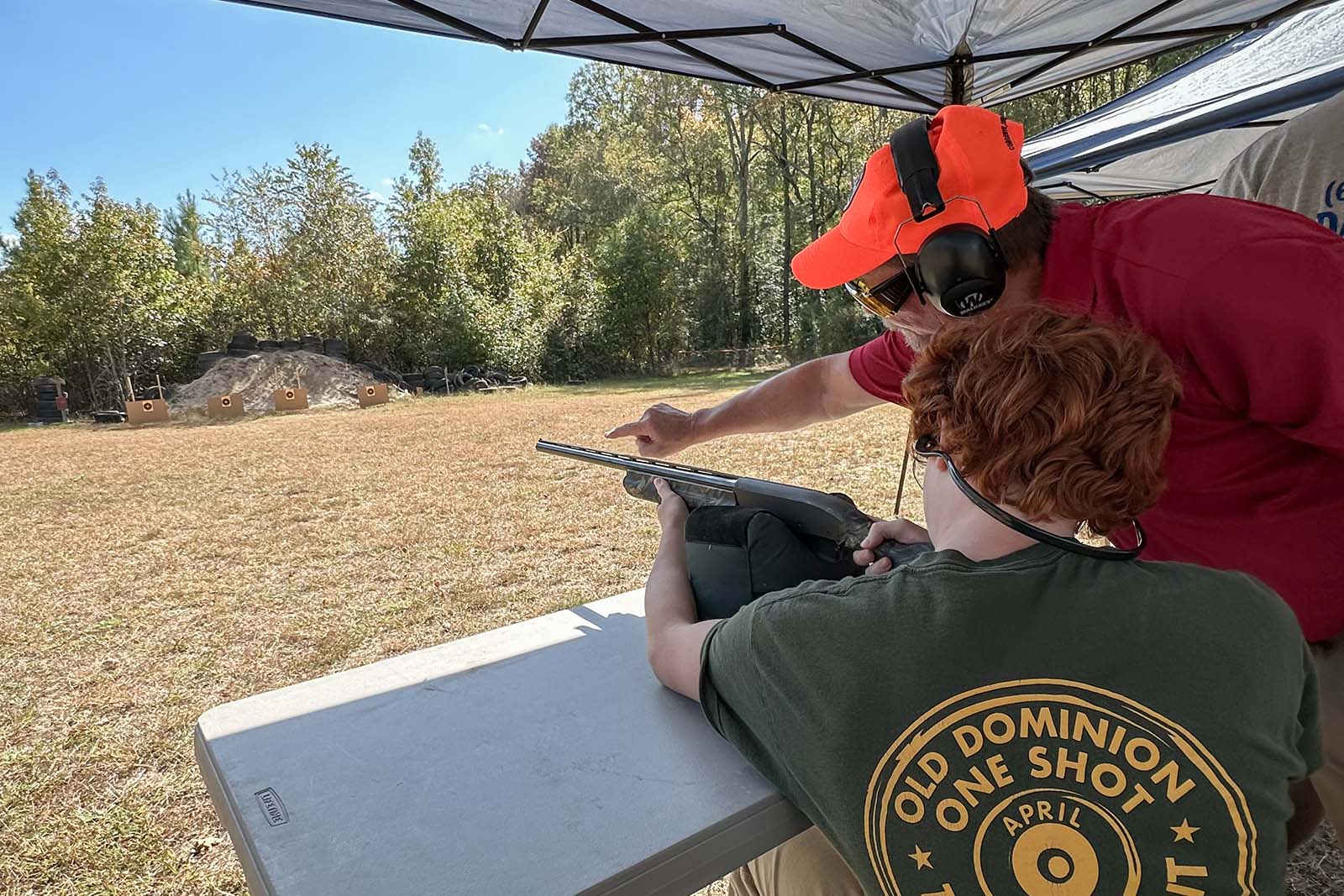 Una persona que practica el tiro al blanco con una escopeta; Un instructor se para cerca señalando el objetivo