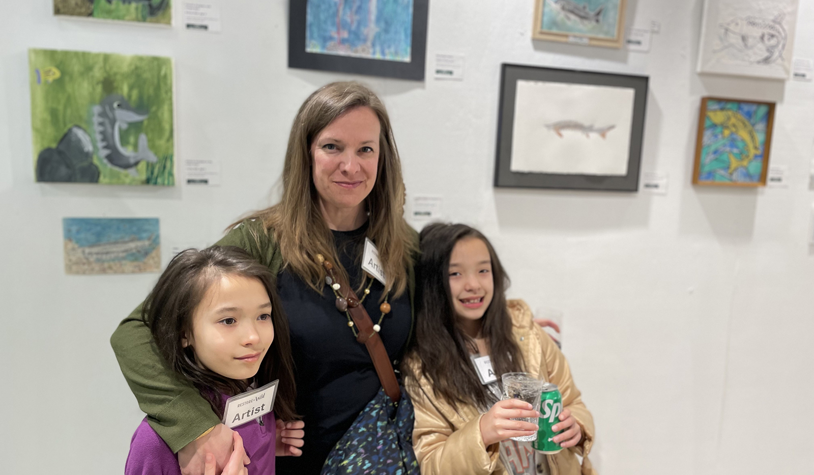 Una foto de una mujer y dos niñas de 8años de pie frente a una pared llena de cuadros.