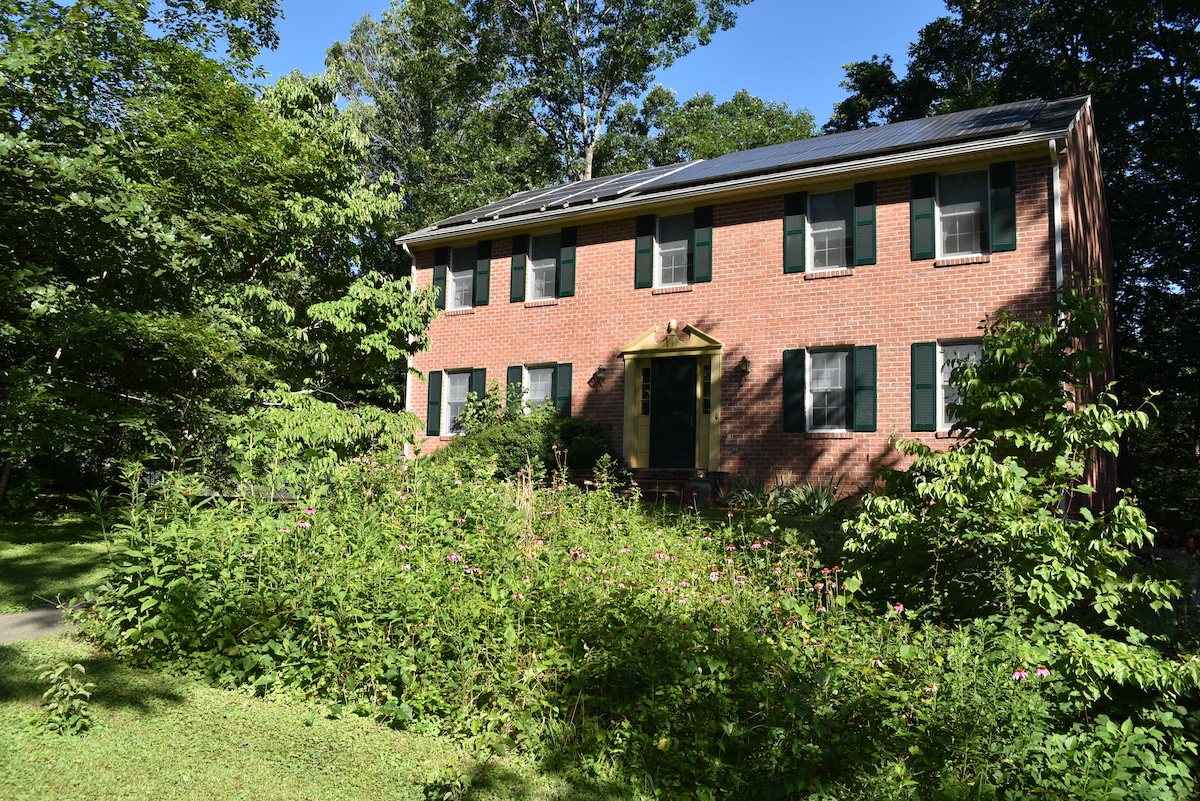 Una imagen de una casa de ladrillo con paneles solares en el techo y un jardín de polinizadores en el patio delantero