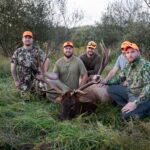 Cinco hombres posando con un alce muerto, rodeados de árboles escasos.