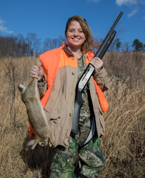 Mindy Tucker con un chaleco de visibilidad naranja con un rifle y un conejo muerto
