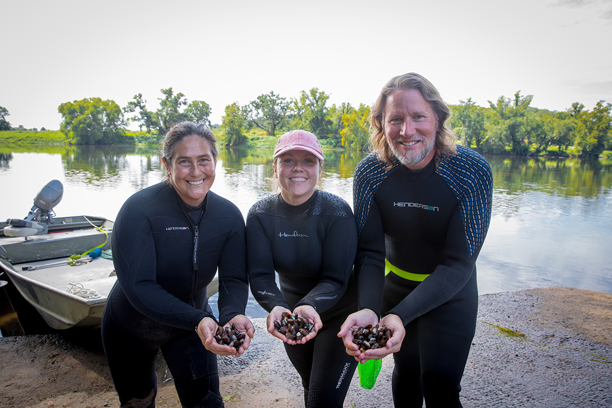 Una imagen de tres empleados de DWR sosteniendo puñados de mejillones con un bote y el río James en el fondo
