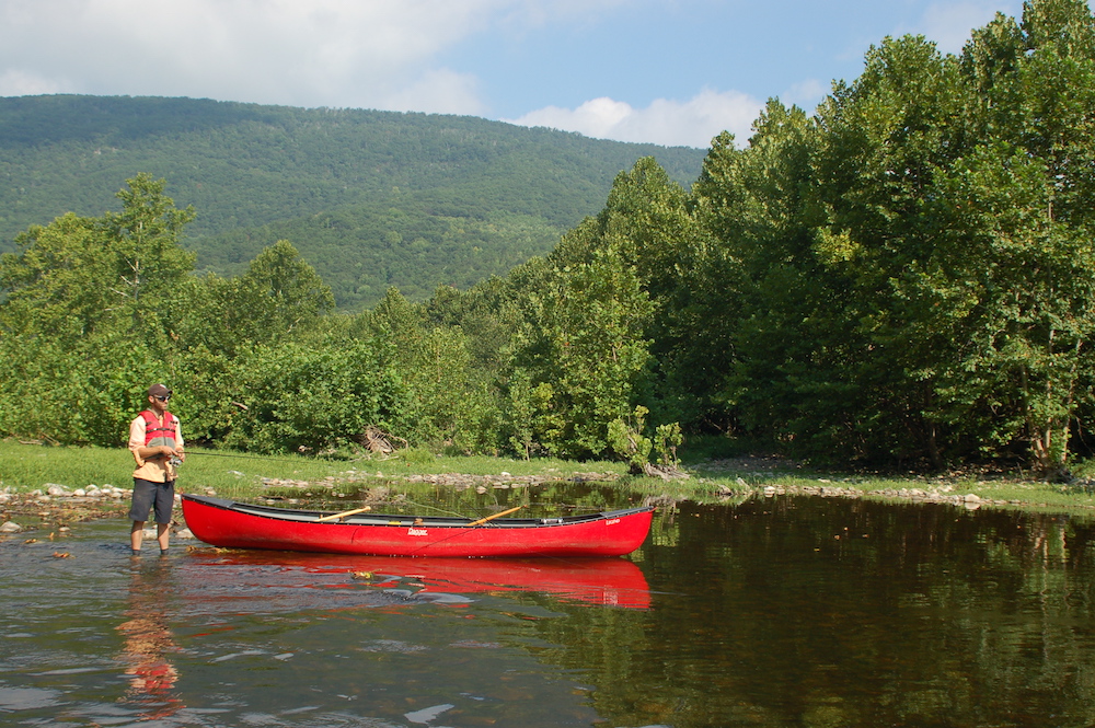 Una imagen de un hombre pescando con un chaleco salvavidas rojo junto a una canoa roja en el río James