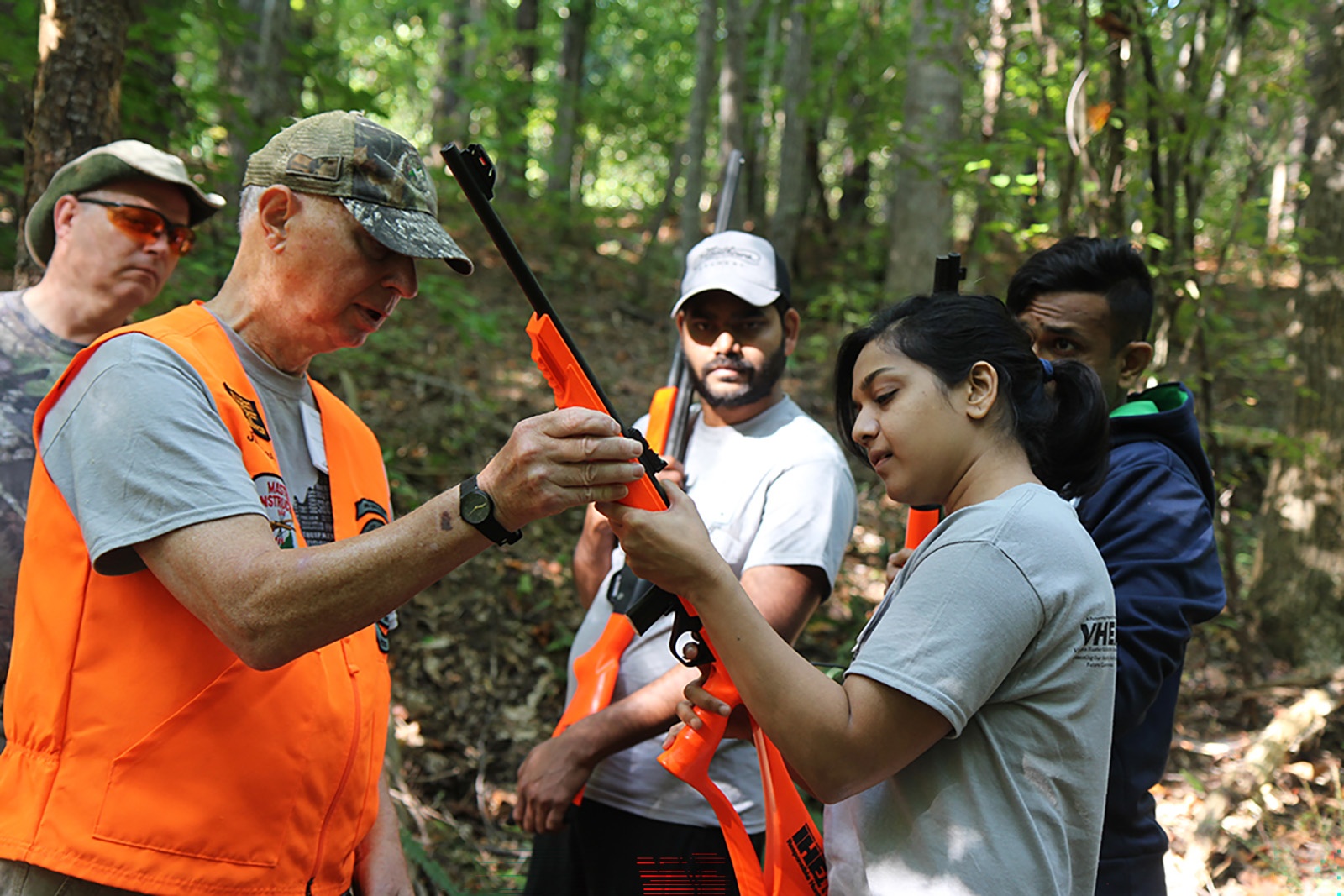 Una fotografía de un hombre explicando a una mujer cómo sostener una escopeta mientras un grupo observa.