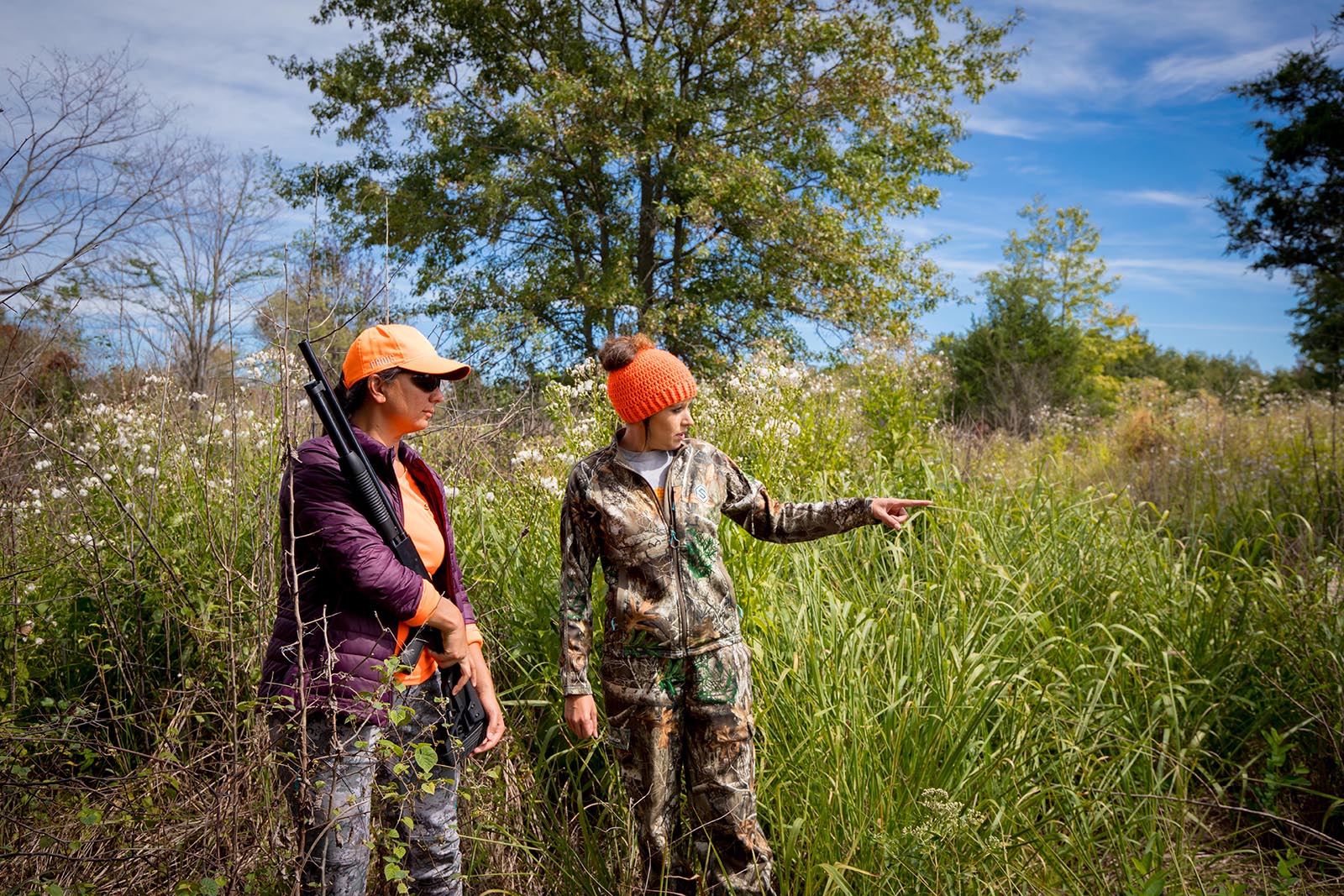 Una foto de dos mujeres con camuflaje y naranja llamativo de pie en un campo, con una mujer dirigiendo a la mujer que sostiene un arma.