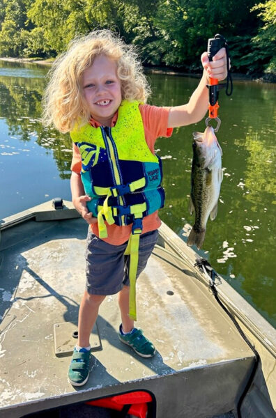Un niño de 5años, con un chaleco salvavidas sonriendo sosteniendo un pez en un anzuelo.