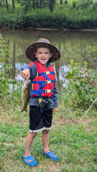 Un niño de 5años, con un sombrero en un chaleco posando, sosteniendo al pez por la boca.