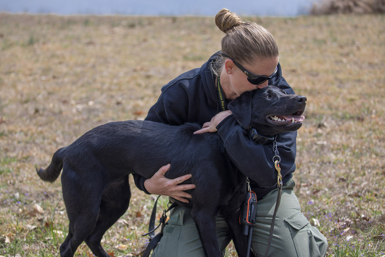 Una foto de una oficial de policía de conservación arrodillada y besando a un perro labrador negro en la cabeza.