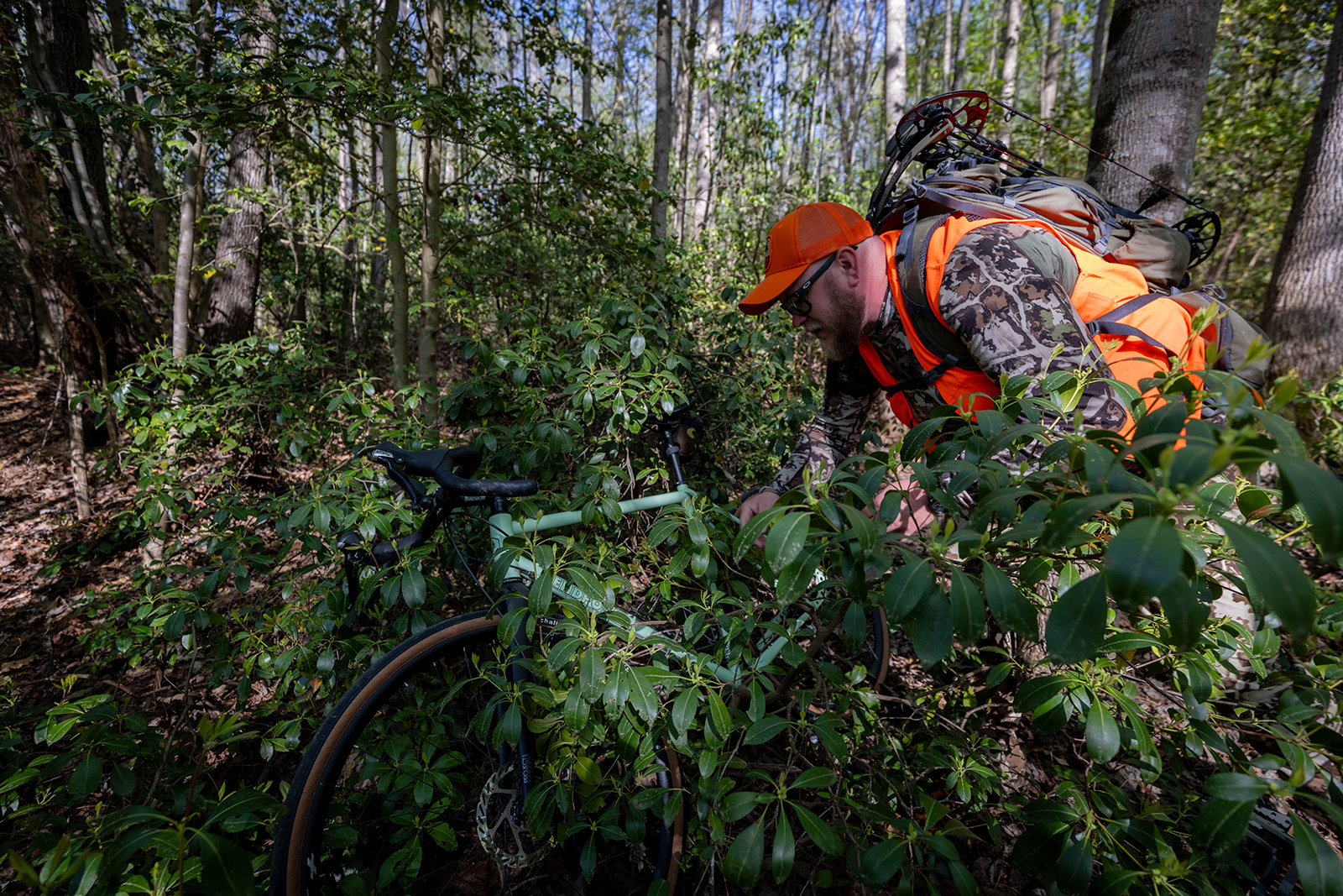 Una foto de un hombre vestido de camuflaje y color naranja brillante cubriendo una bicicleta con arbustos en el bosque.