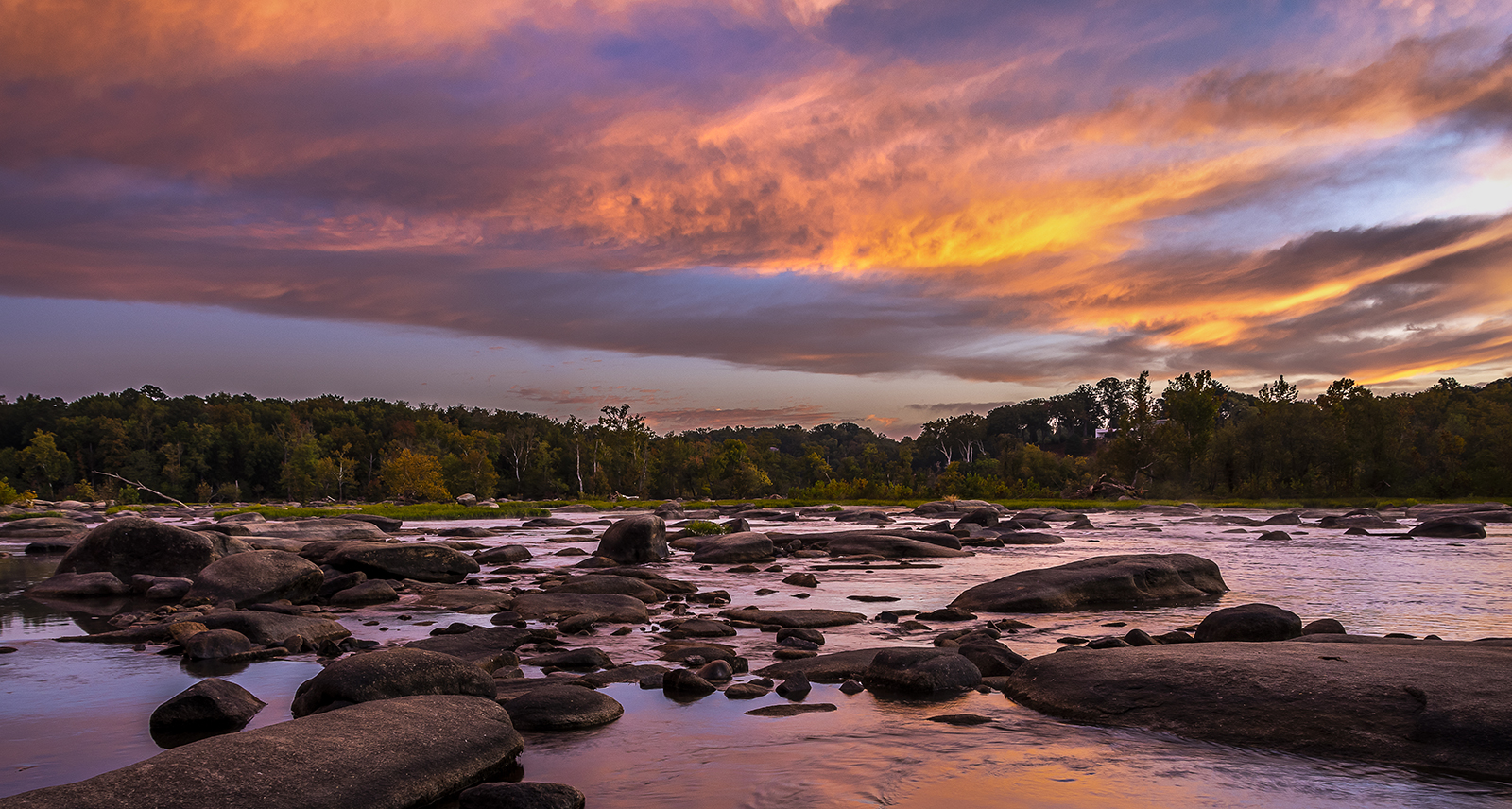 Una foto de un hermoso cielo al amanecer de color naranja, azul y rosa sobre el agua y las rocas de Pony Pasture en el río James en Richmond.