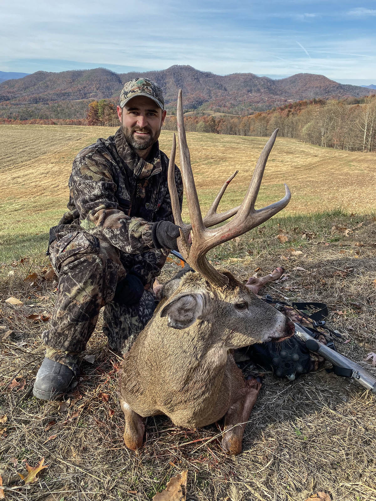 Un cazador vestido de camuflaje posando con un gran ciervo de cola blanca con grandes astas.