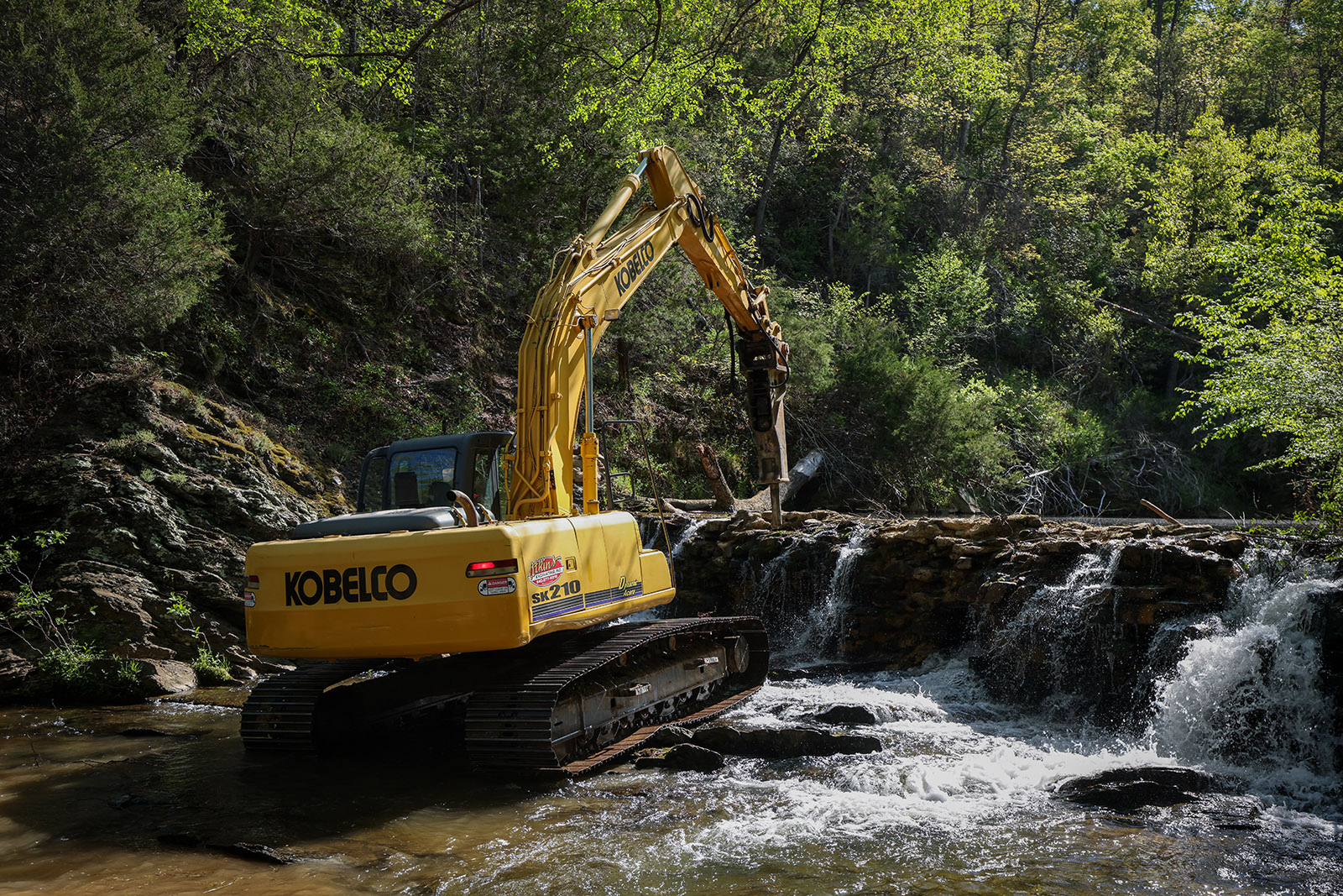 Una foto de una excavadora derribando una vieja presa a través de un pequeño río.