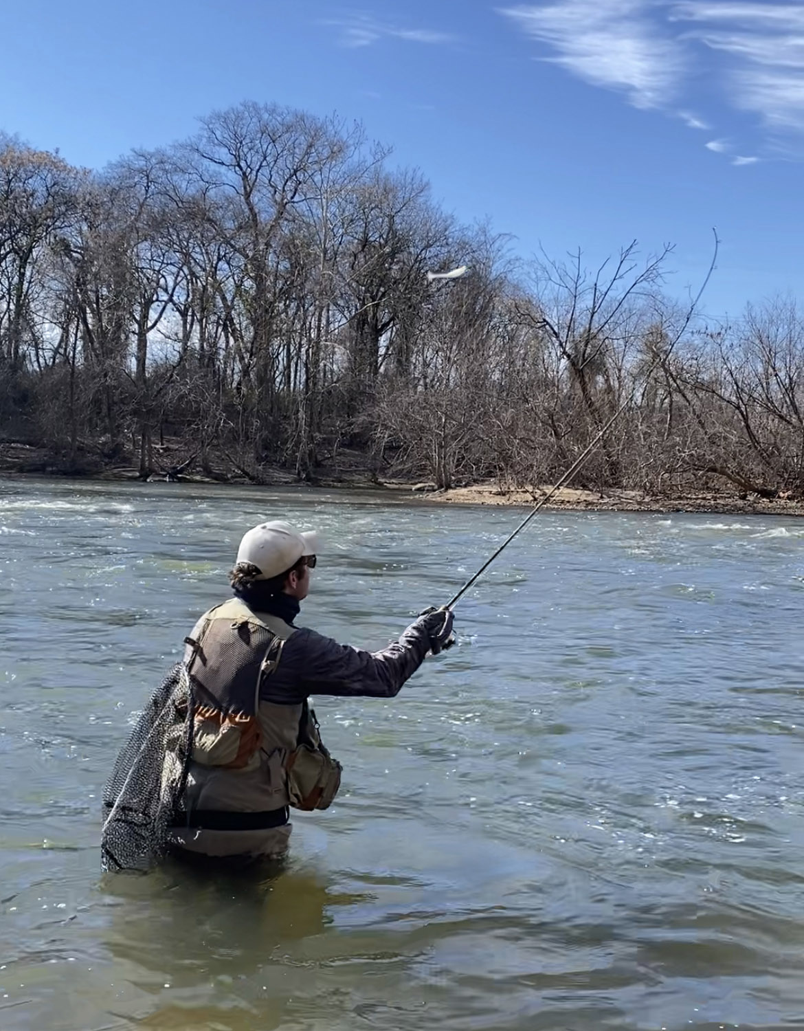 Una foto de un pescador parado hasta las caderas en un río, lanzando una caña giratoria.