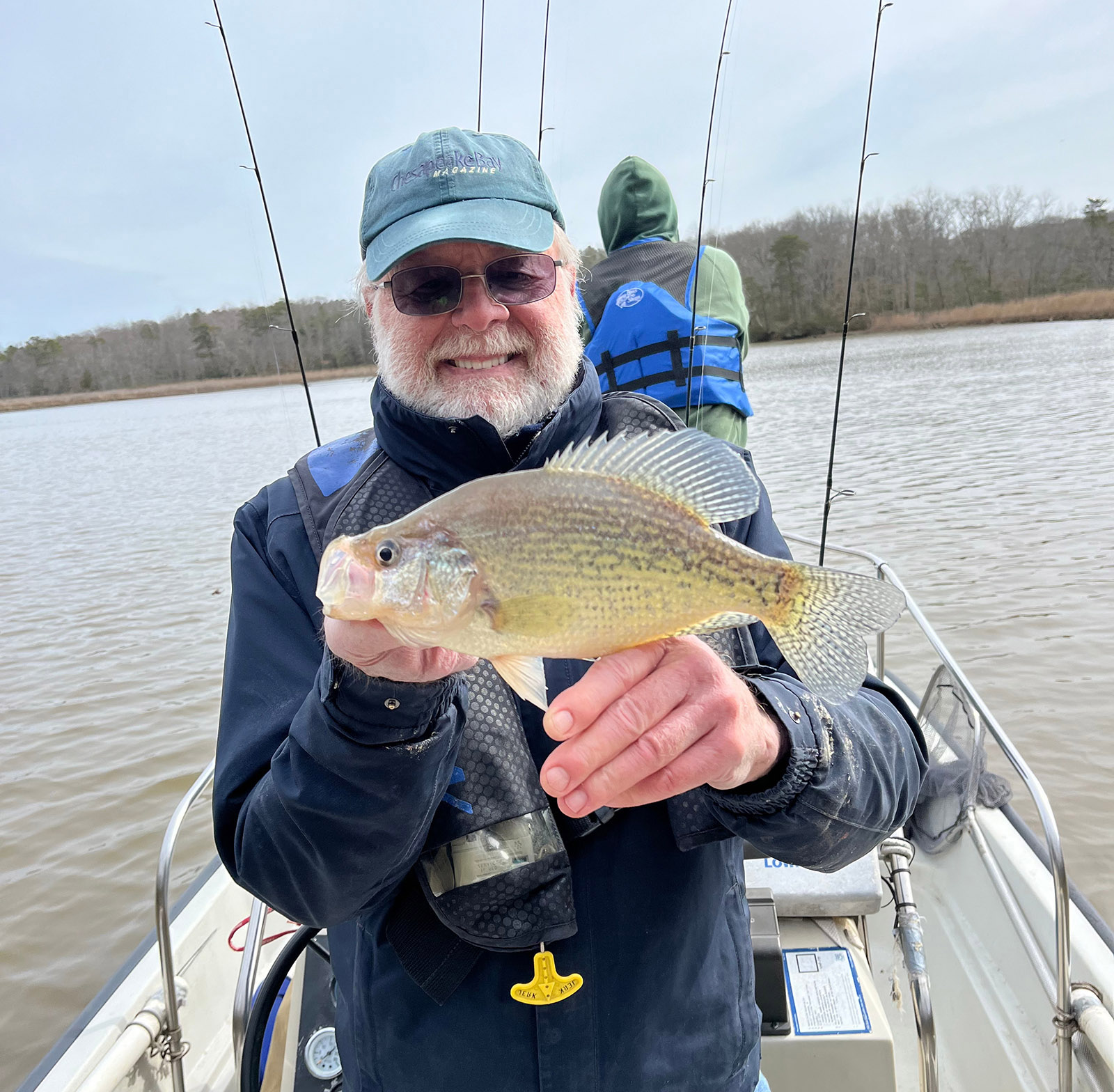 Una foto de un hombre en un bote con un chaleco salvavidas y sosteniendo un gran pez negro mientras sonríe.