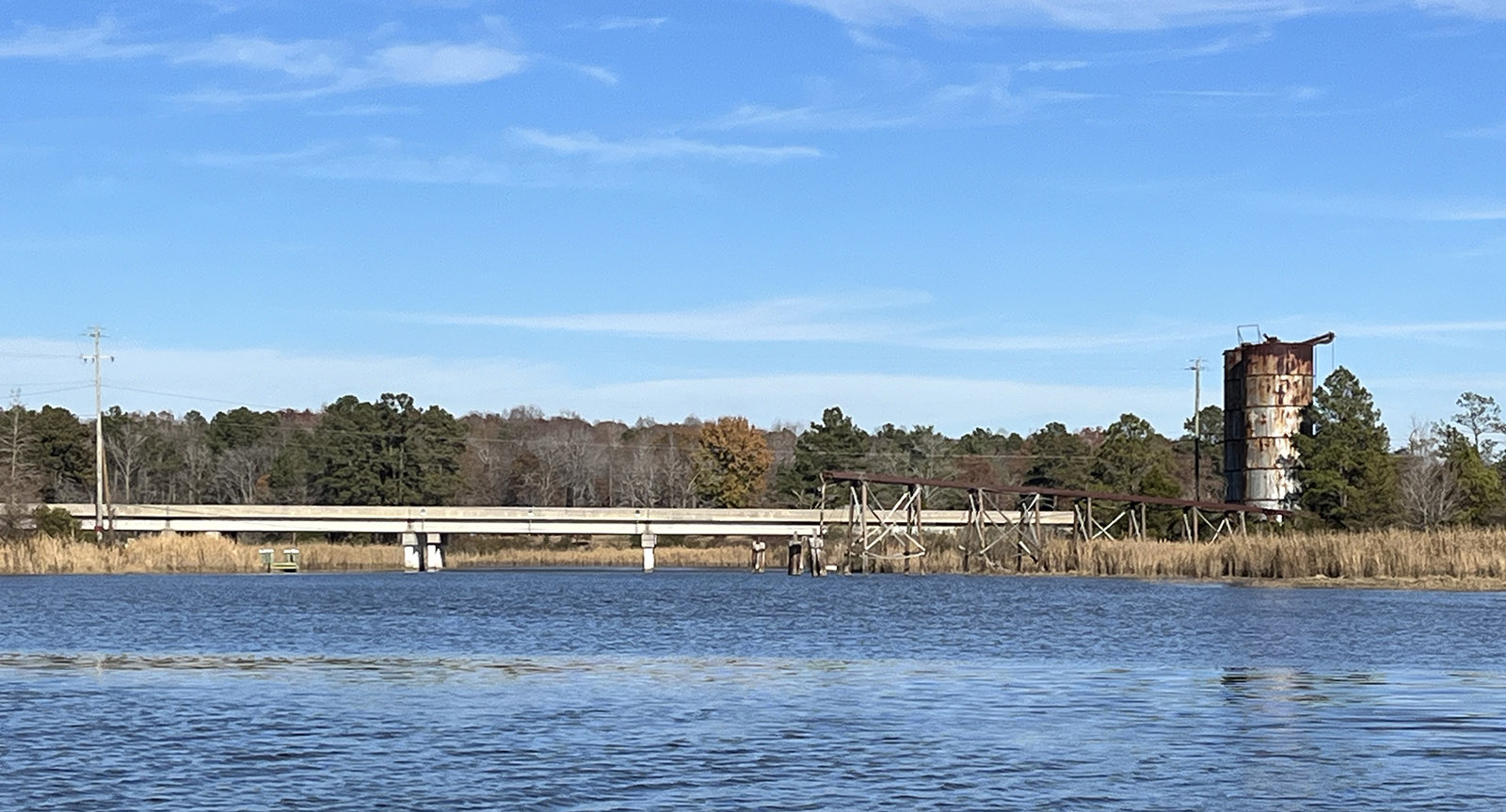 Una foto de un río que fluye bajo un puente bajo de hormigón, con dos silos viejos y ruinosos en la orilla.