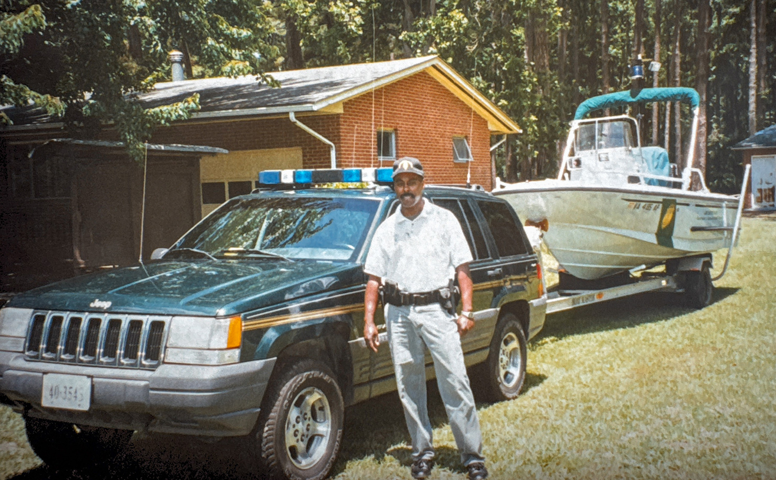 Una foto de un hombre uniformado con pistola y placa de pie frente a un Jeep enganchado a un remolque de barco.