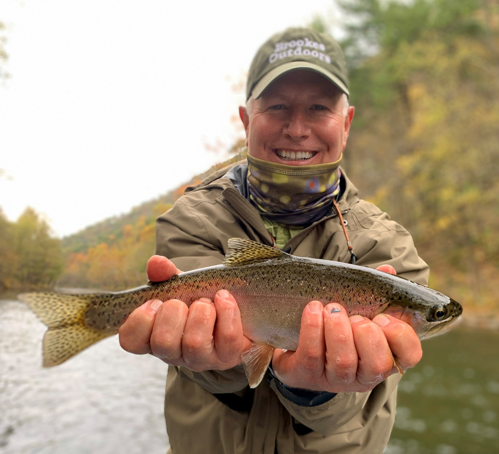 Un hombre de pie en un río, sosteniendo una gran trucha arco iris con ambas manos.