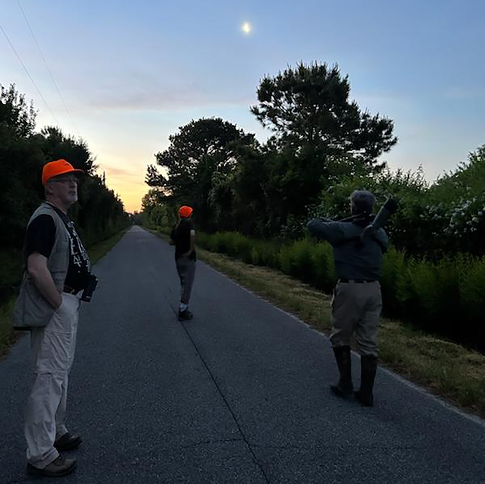 Una foto de tres personas caminando por una carretera en un área remota a la luz del amanecer con la luna en el cielo.