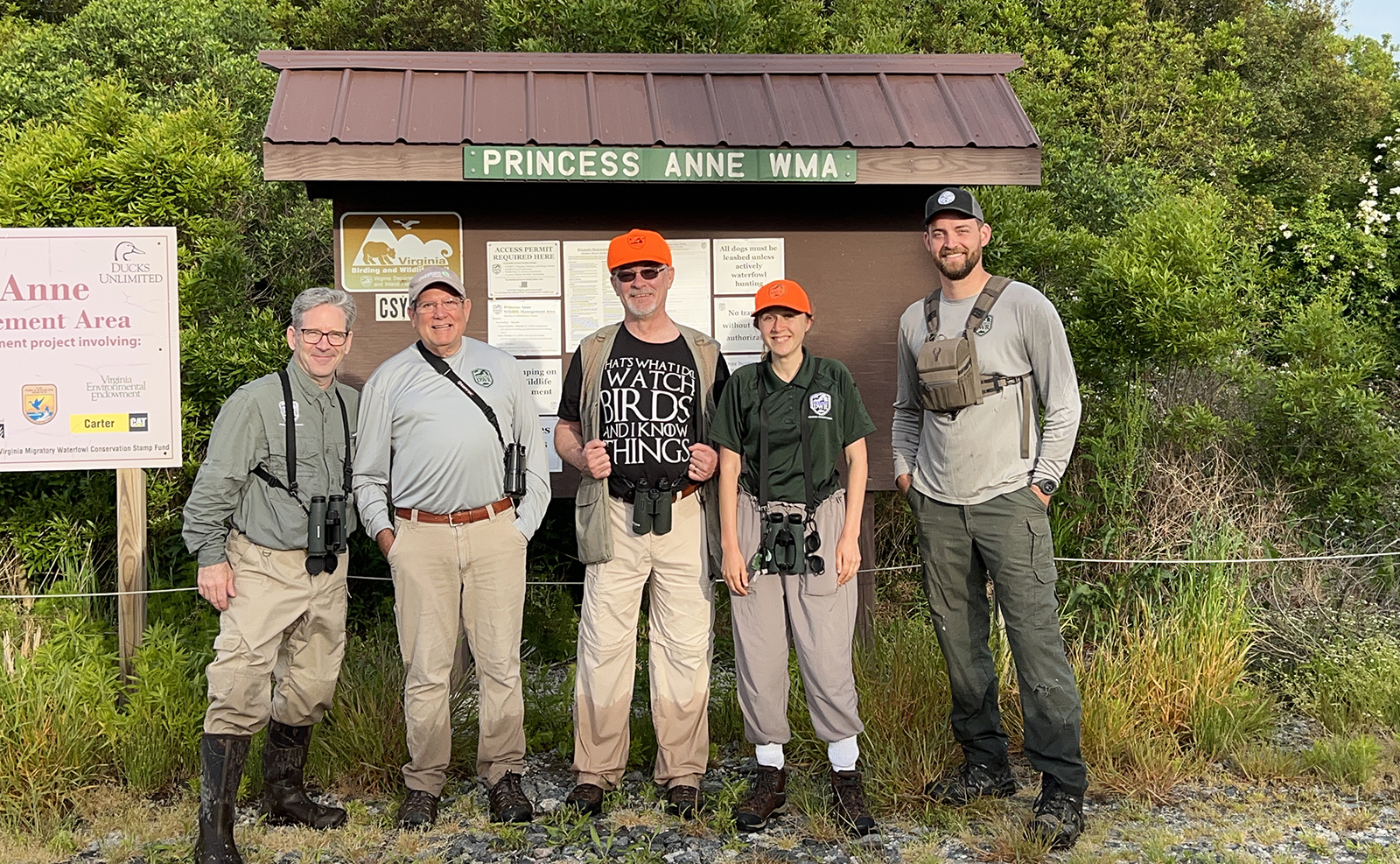 Un grupo de cinco personas con binoculares posando frente a un quiosco de letreros con la etiqueta Princesa Ana WMA.