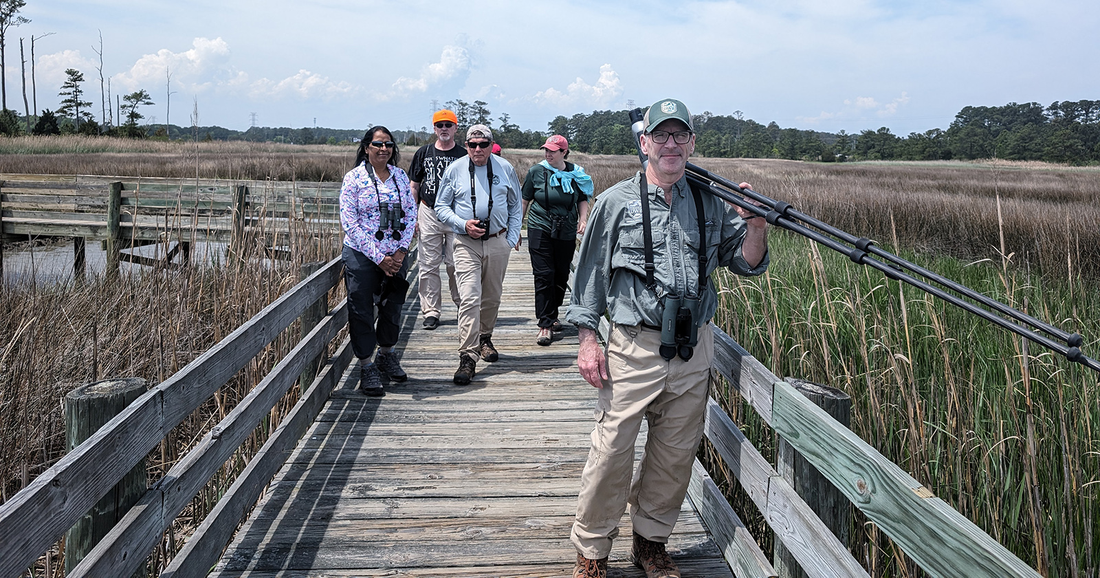Una foto de cinco personas con binoculares caminando por una pasarela de madera a través de un humedal.