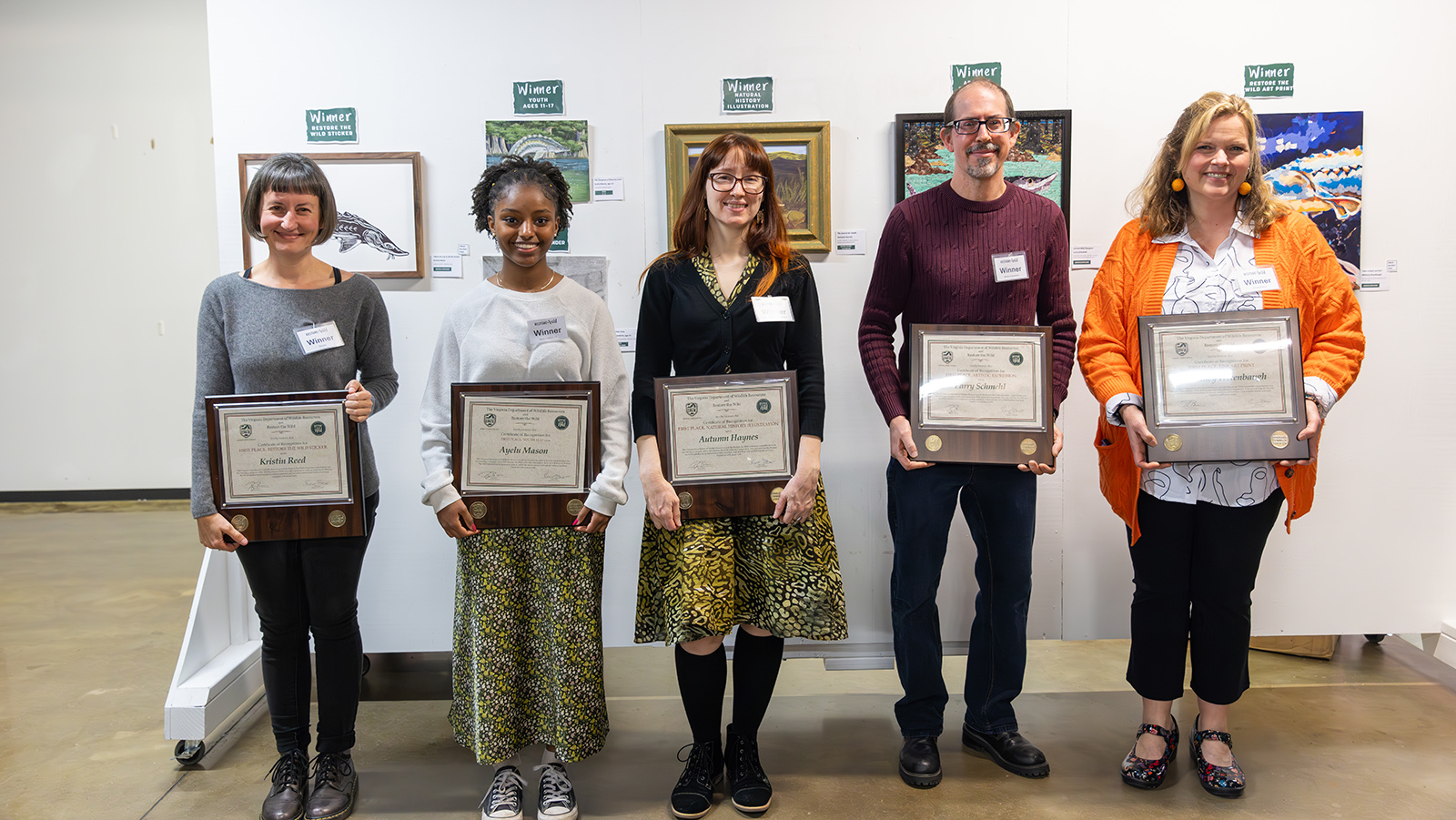 Una foto de cinco personas de pie sosteniendo certificados frente a una pared con obras de arte.