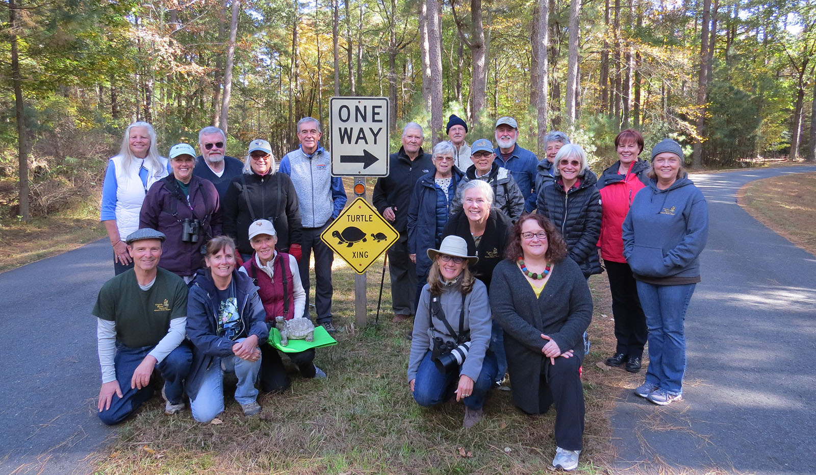 Un grupo de 20 personas se reunió alrededor de un letrero de cruce de tortugas en la mediana de una pequeña carretera pavimentada, con una persona arrodillada al frente con una estatua de tortuga.
