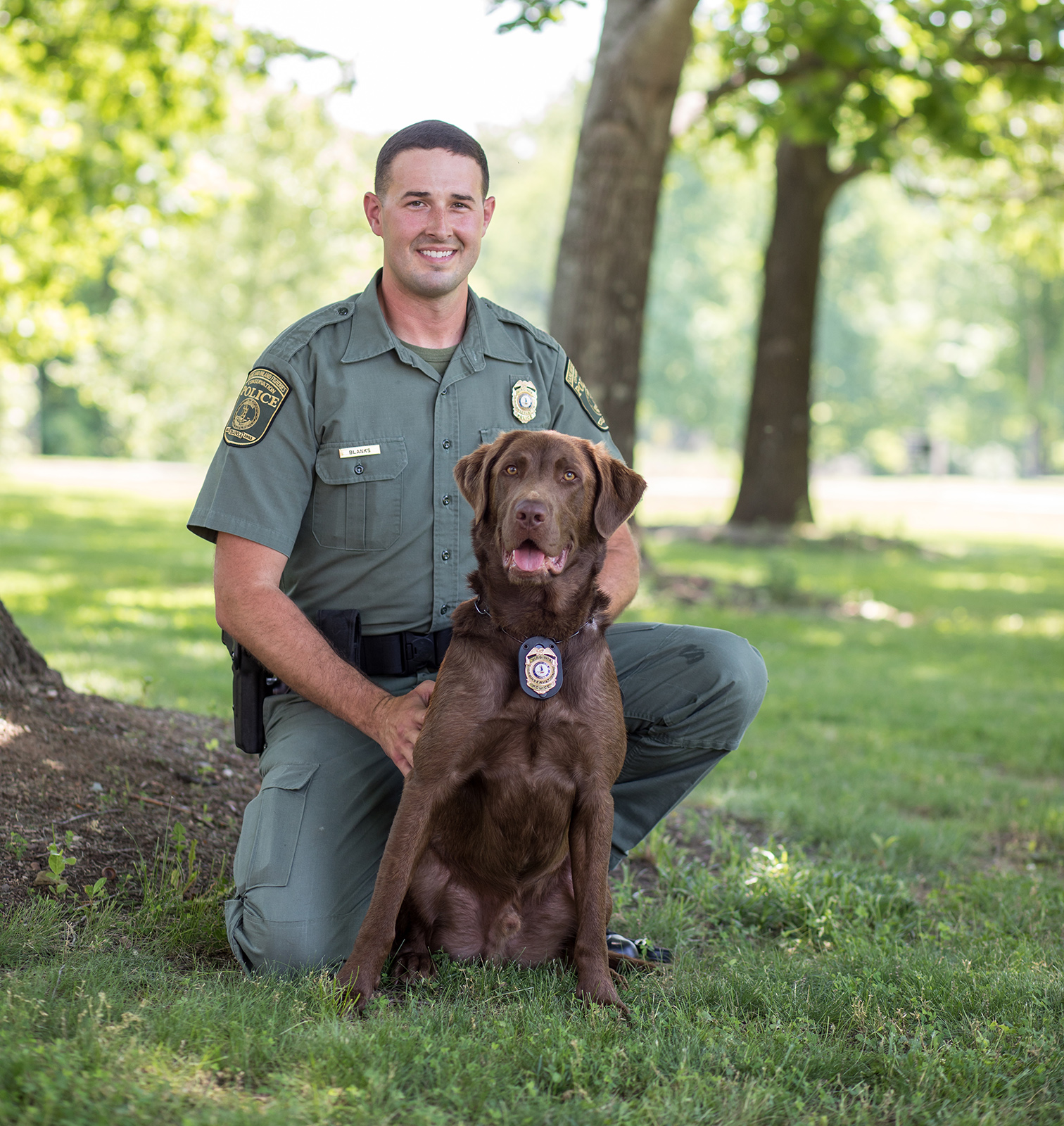 A photo of a man in law enforcement uniform kneeling next to a sitting dog wearing a badge on its collar.