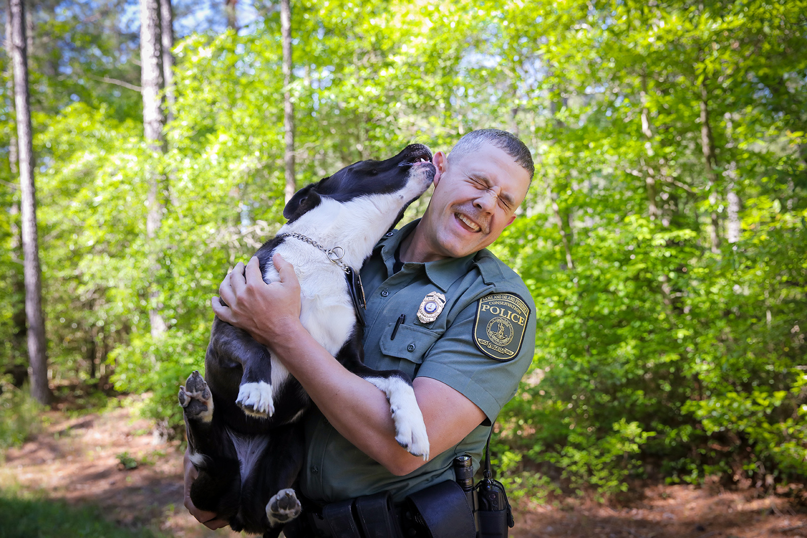 A photo of a man in law enforcement uniform holding a dog and smiling while it licks him on the ear.