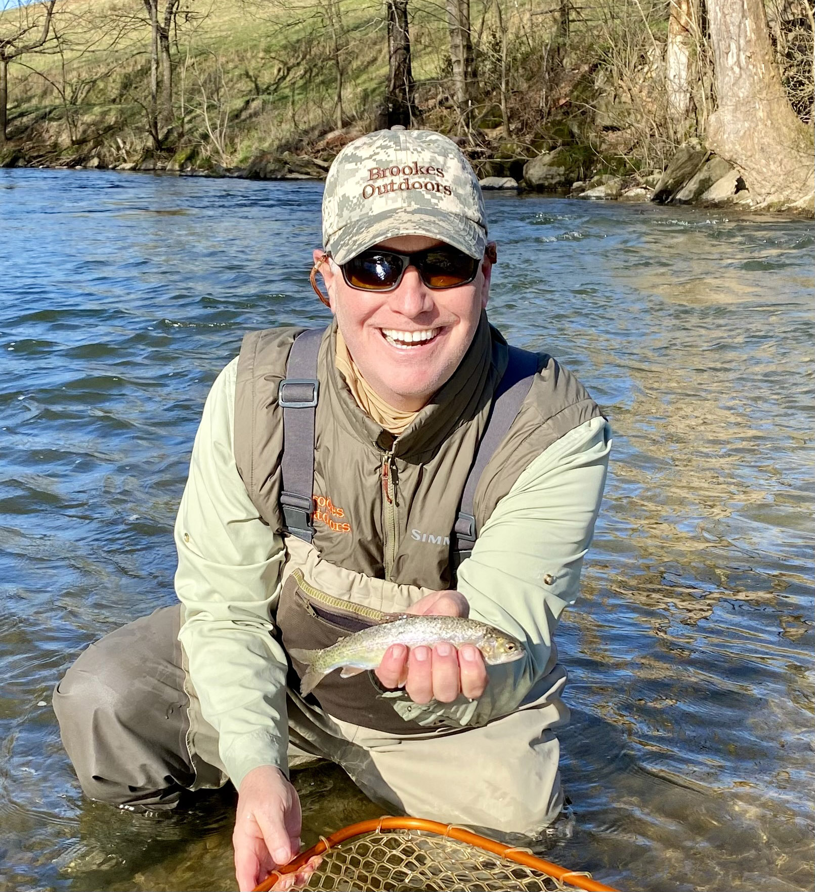 Una foto de un hombre arrodillado en un río, sosteniendo una pequeña trucha arco iris de una red.