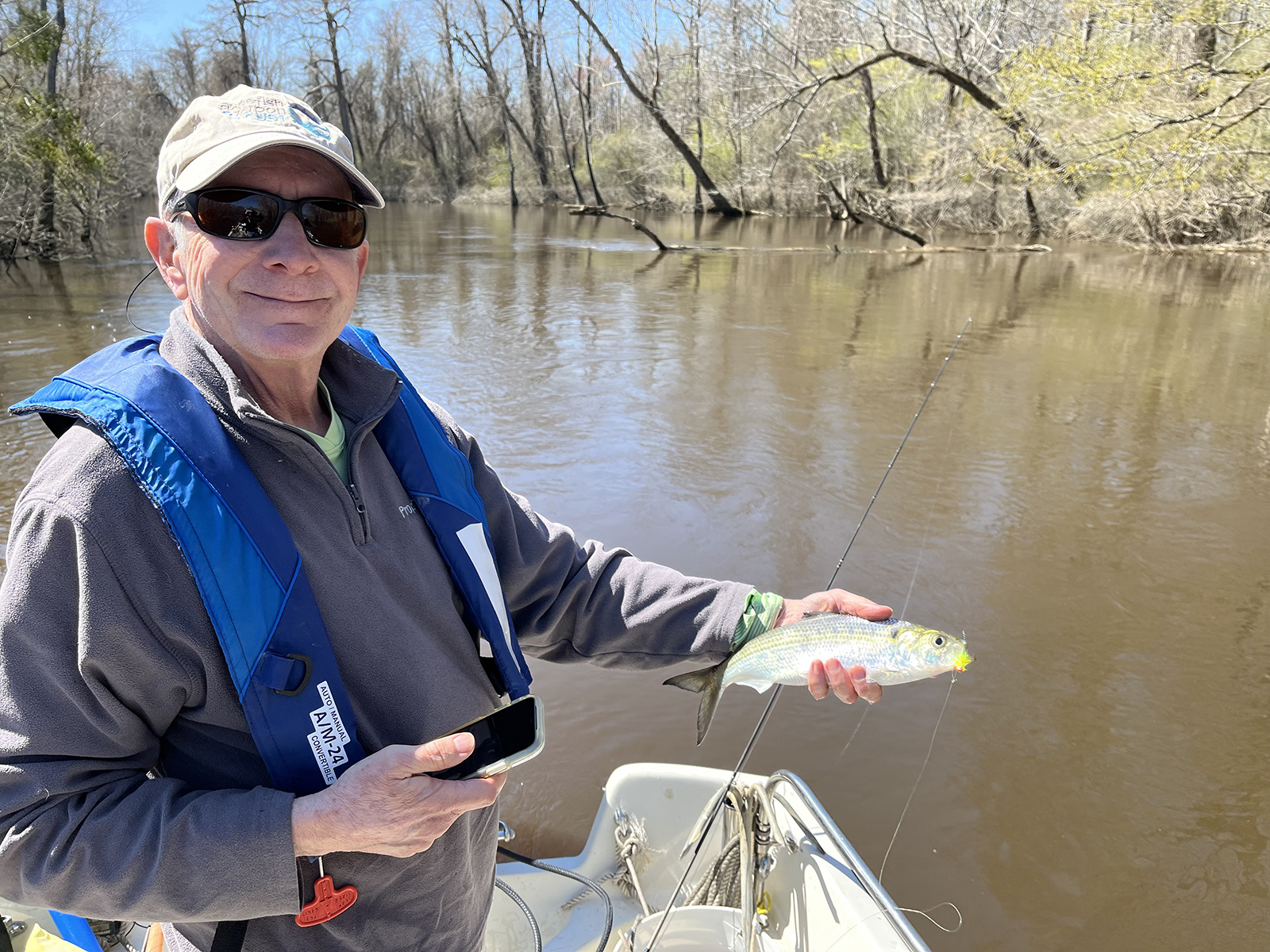 Un hombre con un chaleco salvavidas, de pie en un bote en un río y sosteniendo un pez sábalo brillante.