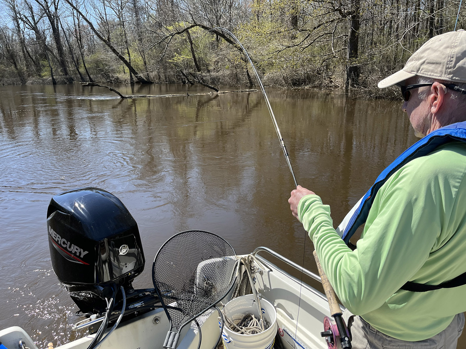 Una foto de un hombre de pie en un bote con una caña de pescar con mosca inclinada sobre el agua.