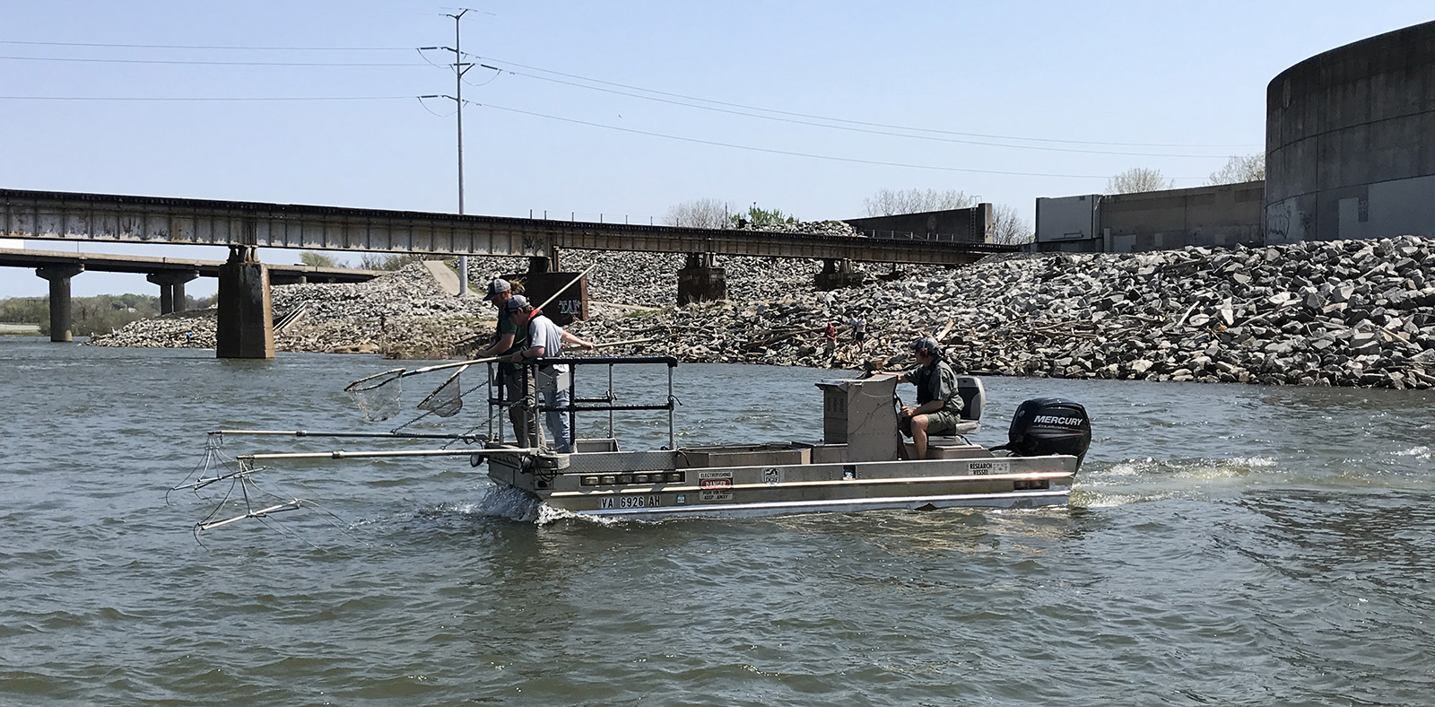 Una foto de dos personas en un bote de metal en un río, con postes que se extienden hacia el agua desde el bote y una persona de pie en la proa con una red.