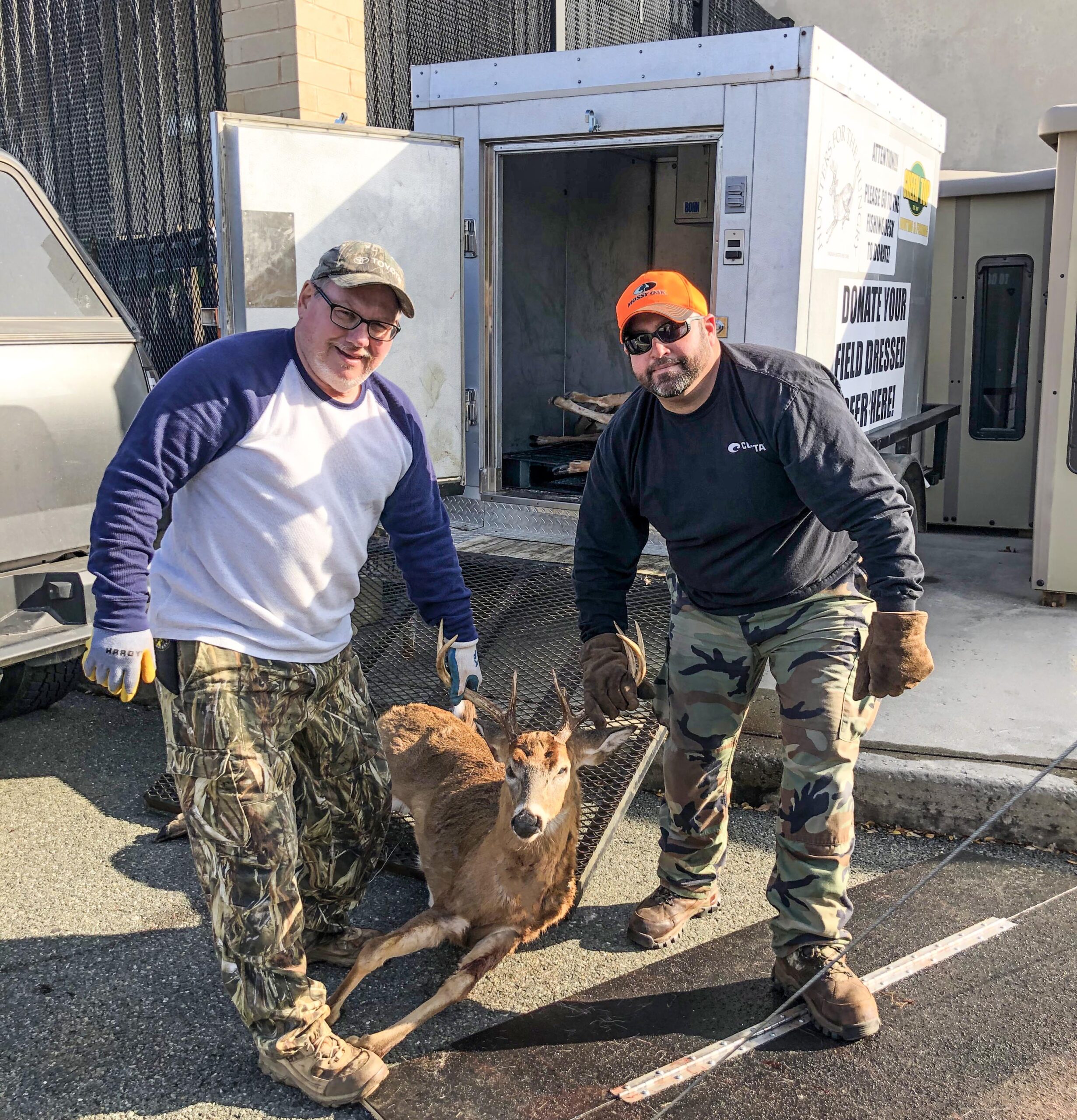 Una foto de dos hombres sosteniendo las astas de un ciervo cazado frente a un remolque refrigerado.