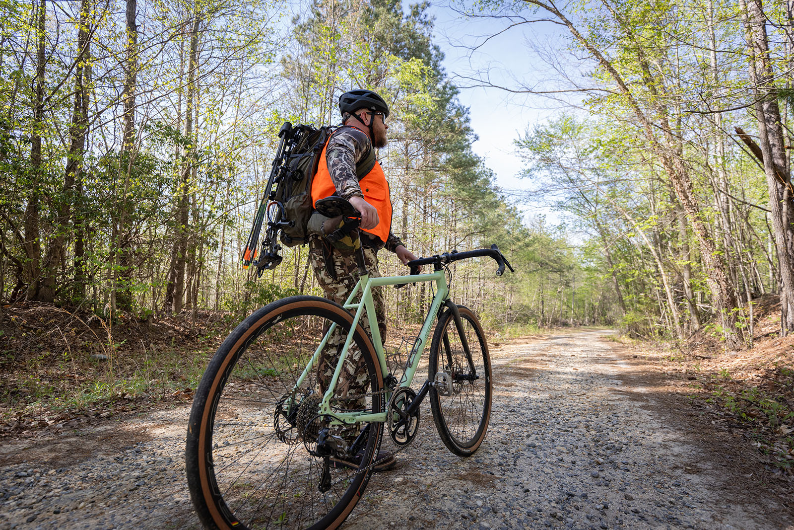 Una foto de un hombre vestido de camuflaje y color naranja brillante, con un casco de bicicleta y una mochila grande, se encuentra junto a una bicicleta en un camino de grava en el bosque.