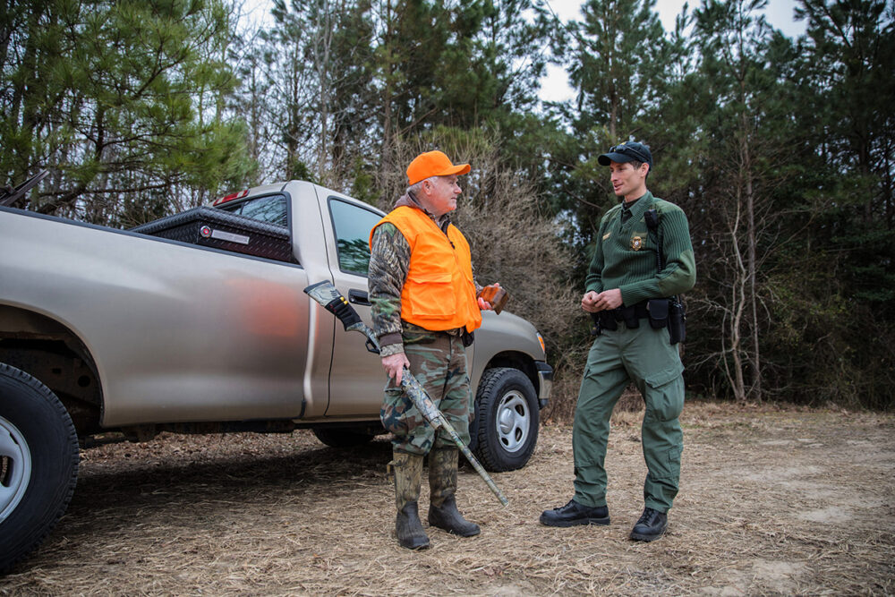 Una imagen de un cazador con un camión y un chaleco de visibilidad naranja hablando con un hombre vestido de verde que es un oficial de policía de conservación