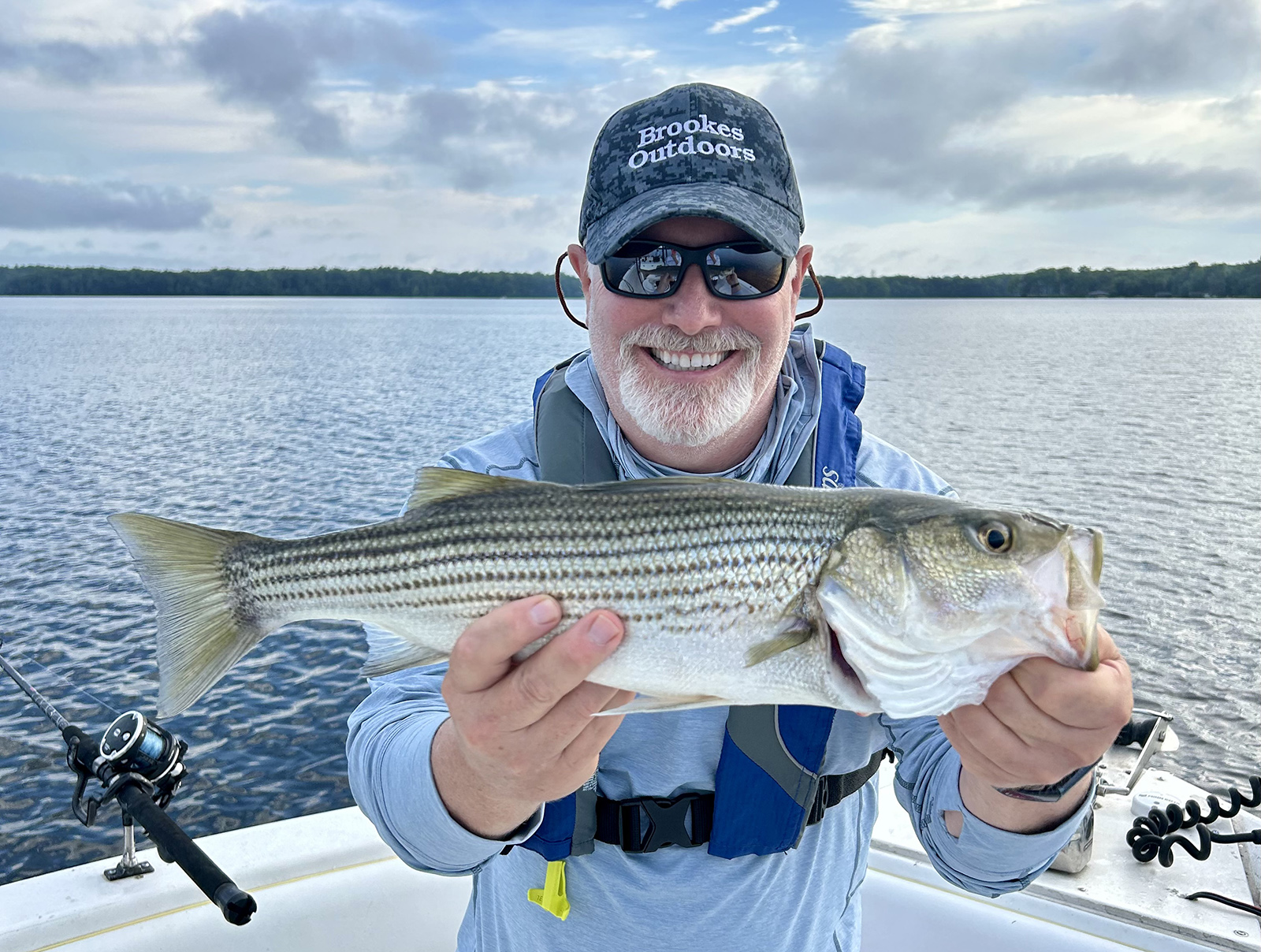 Una foto de un hombre sonriente con un chaleco salvavidas y sosteniendo una gran lubina rayada en un bote con un lago al fondo.
