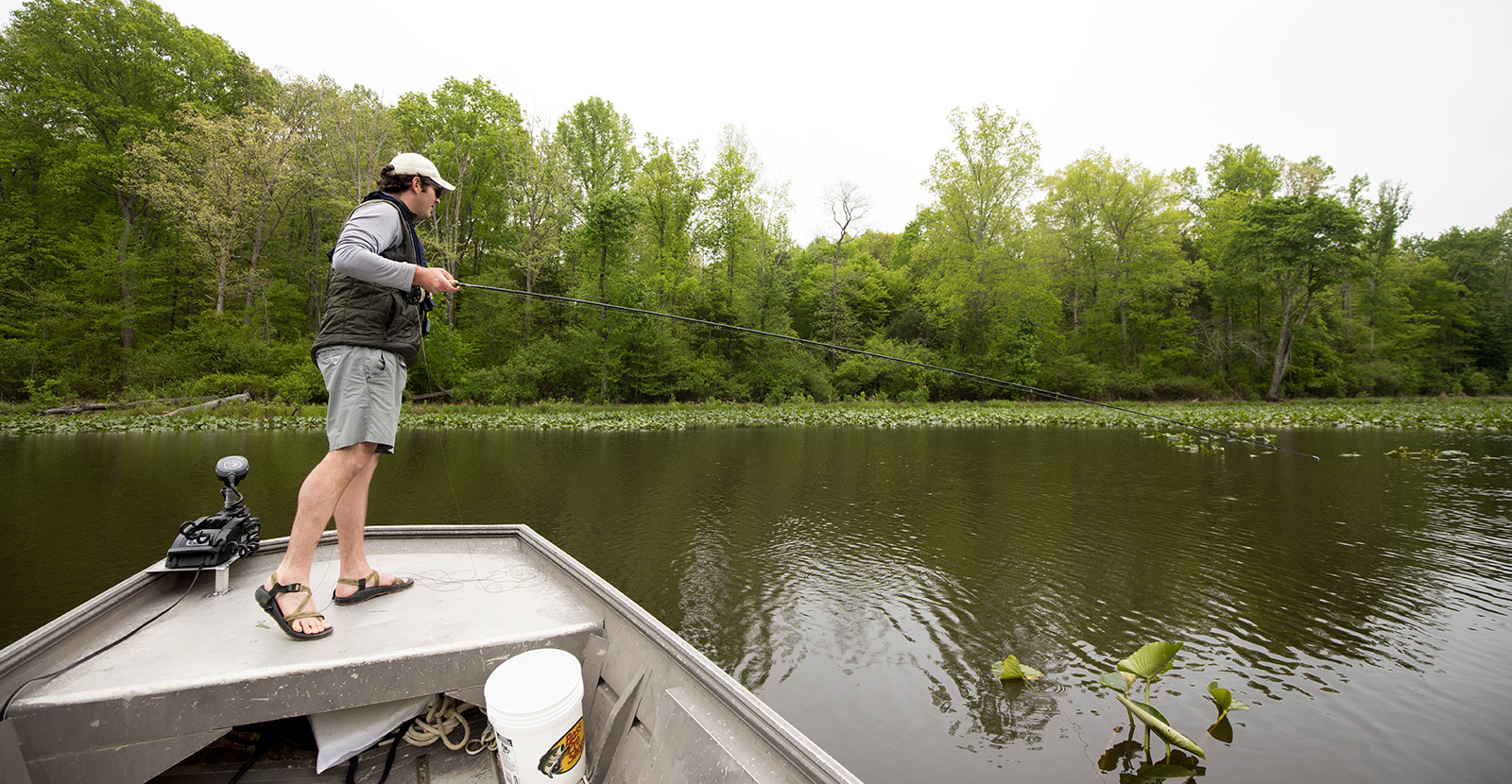 Un hombre de pie en la proa de un bote, sosteniendo una caña de pescar y mirando al agua.