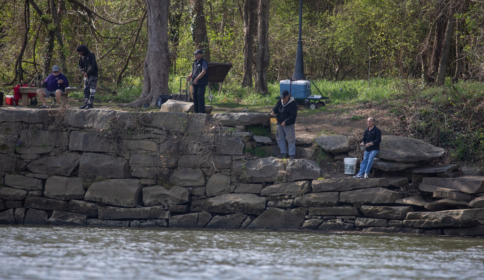 Una foto de personas de pie y sentadas en la orilla del río pescando.