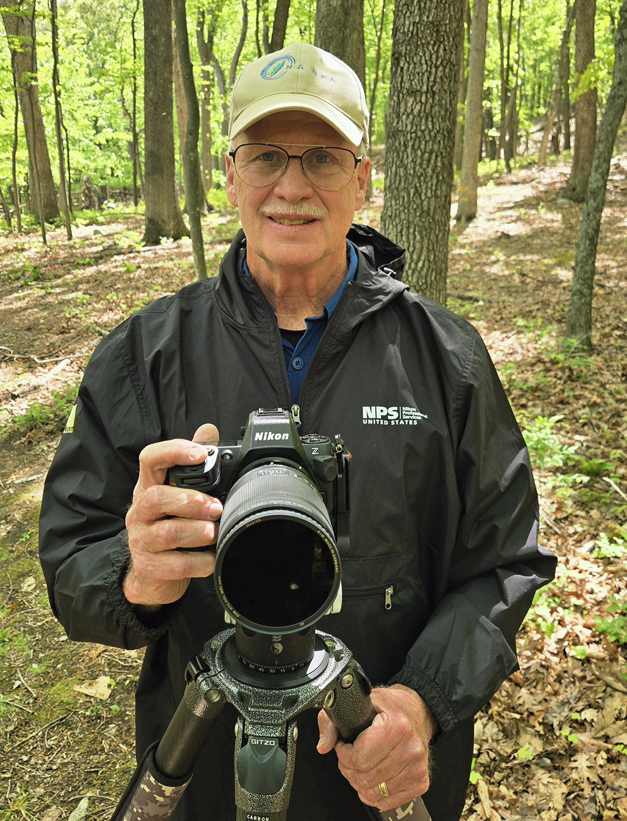 Una foto de un hombre sosteniendo una cámara conectada a un trípode mientras está de pie en el bosque.