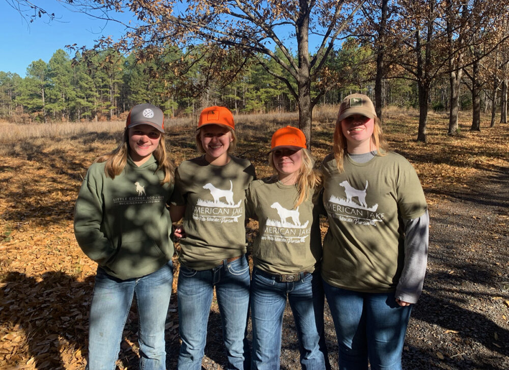 Una imagen de cuatro adolescentes en un bosque caducifolio posando para una foto; con camisetas que dicen "American Jake" que tienen un perro en ellas