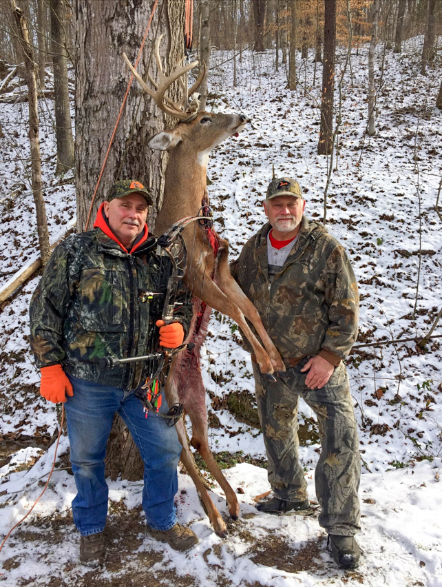 Una foto de dos hombres en el bosque con nieve en el suelo, de pie junto al cuerpo de un gran ciervo de cola blanca.