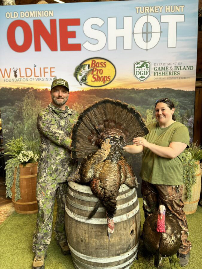 Una foto de un hombre y una mujer de pie frente a un telón de fondo con One Shot y los nombres de los patrocinadores con un pavo grande exhibido.